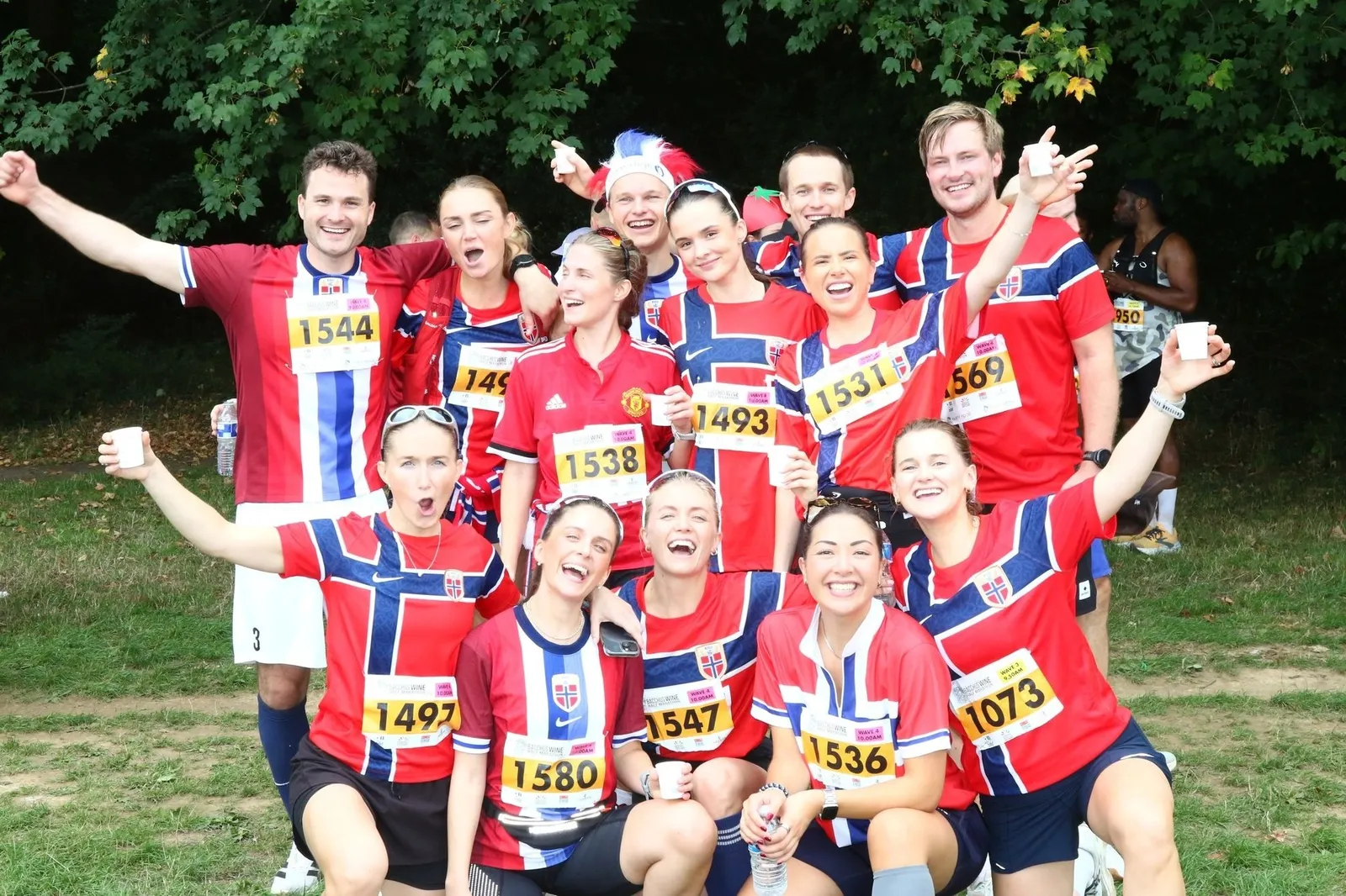 A group of smiling runners in red, white and blue Norwegian shirts and race numbers raising cups at the Bacchus Run