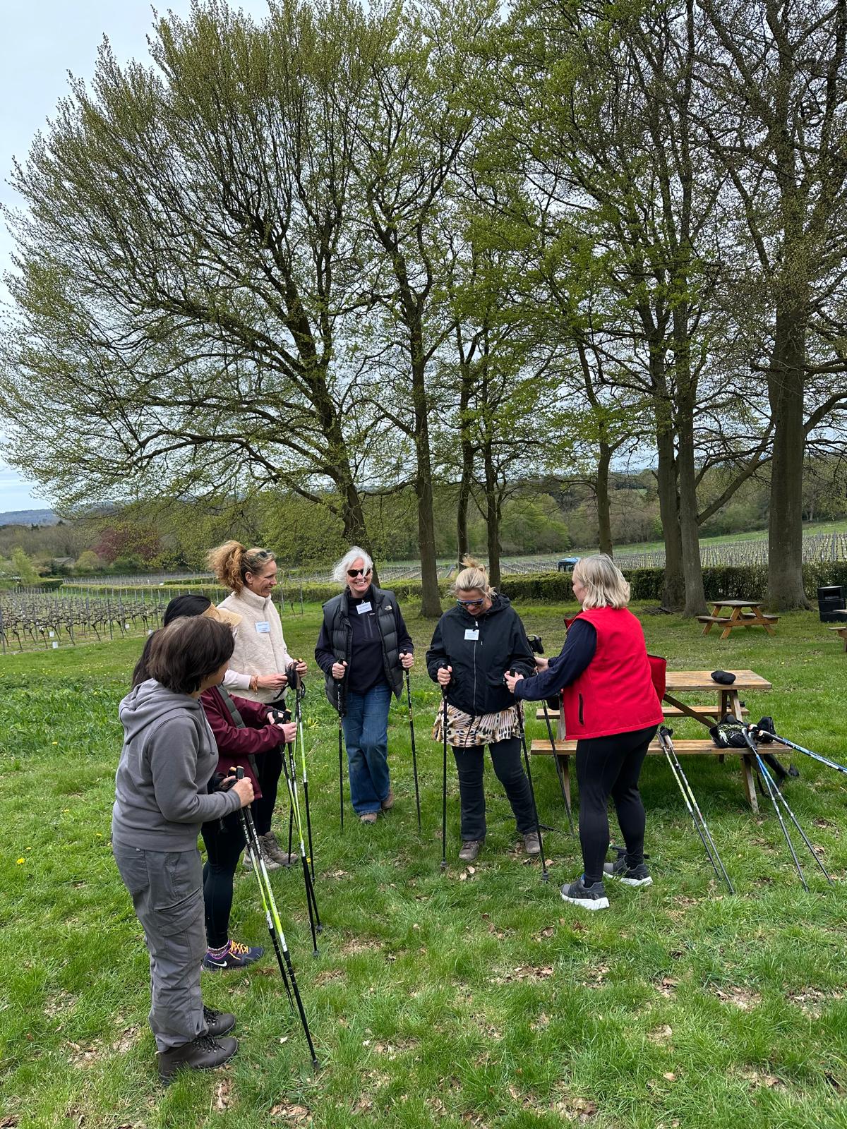 Members with walking poles gathered under trees beside a vineyard before a Nordic walking networking session